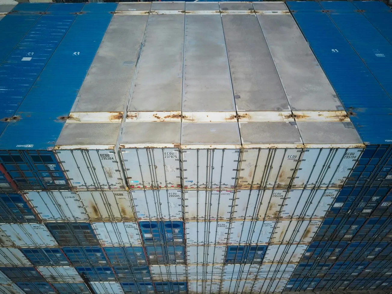 Overhead view of stacked shipping containers in varying shades of blue and gray, showing signs of rust. Industrial and geometric patterns dominate.
