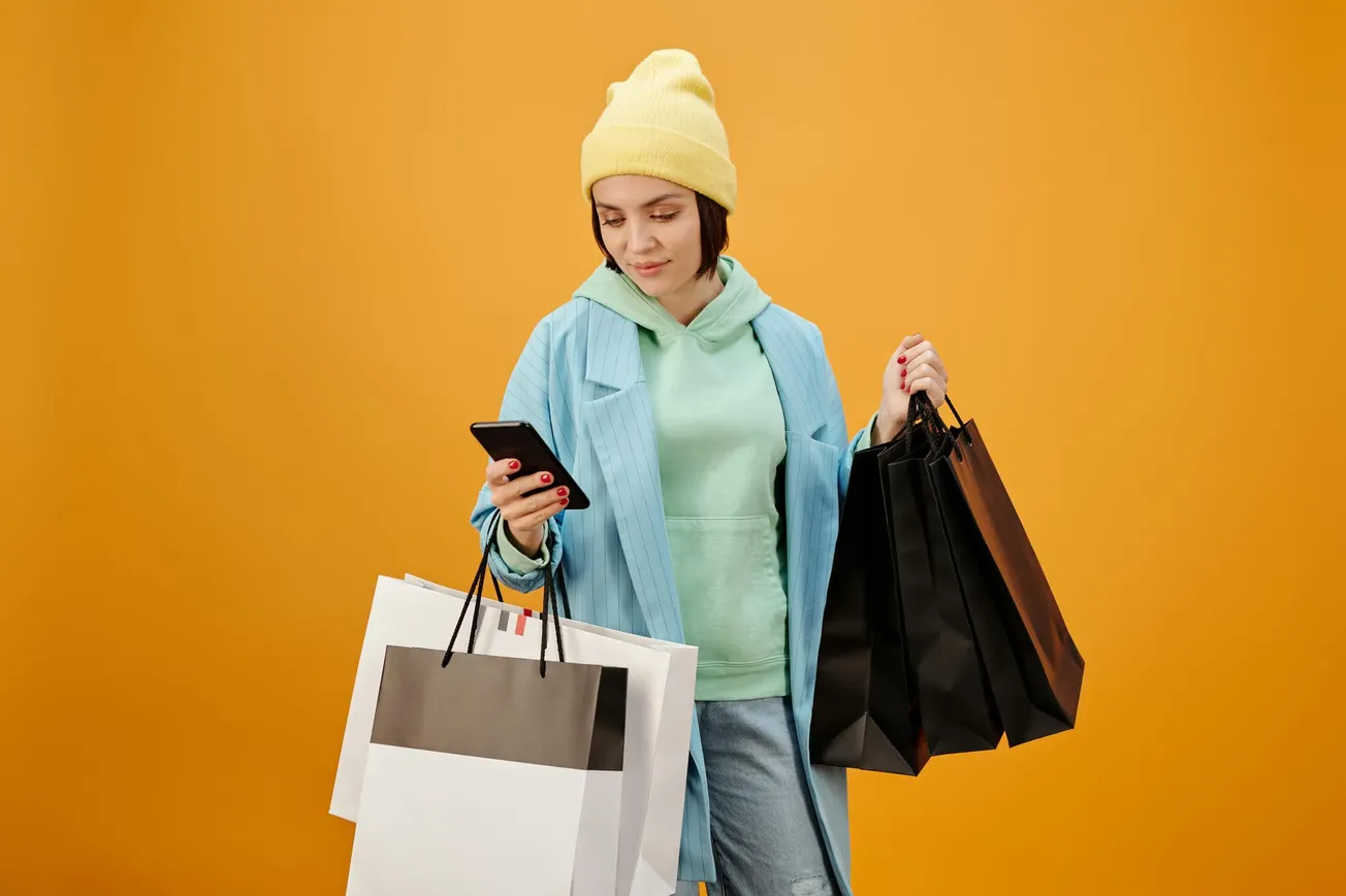 Young woman holding shopping bags and checking her phone after retail shopping against an orange background.