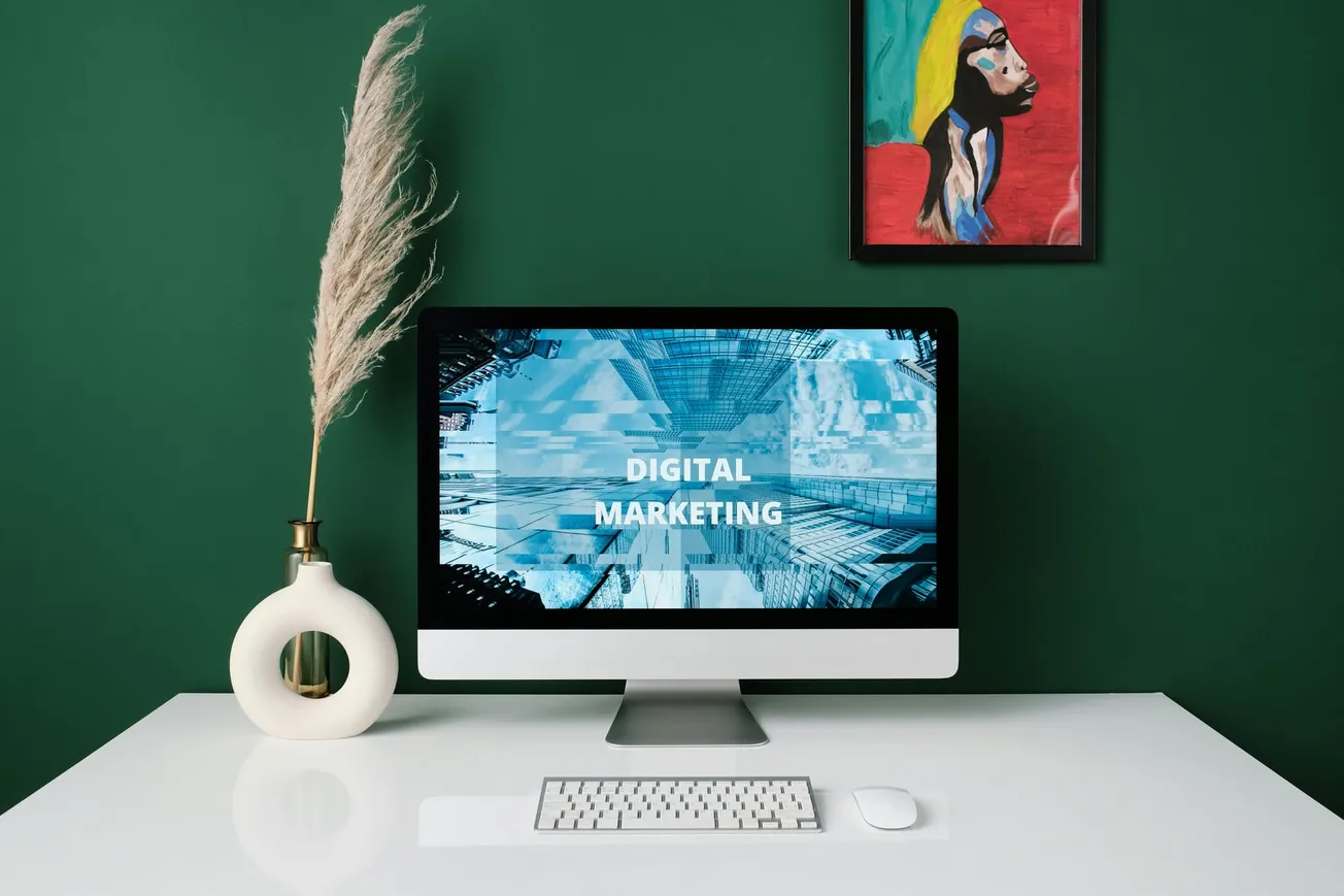 A computer on a white desk displays "Digital Marketing" on the screen. A minimalist vase with pampas grass is beside it. A colorful portrait hangs on a dark green wall.