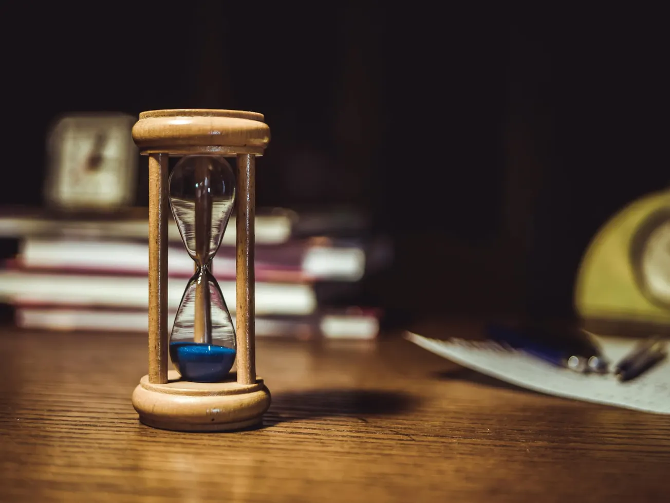 A wooden hourglass with blue sand is on a wooden table, surrounded by closed books and pens under soft lighting.