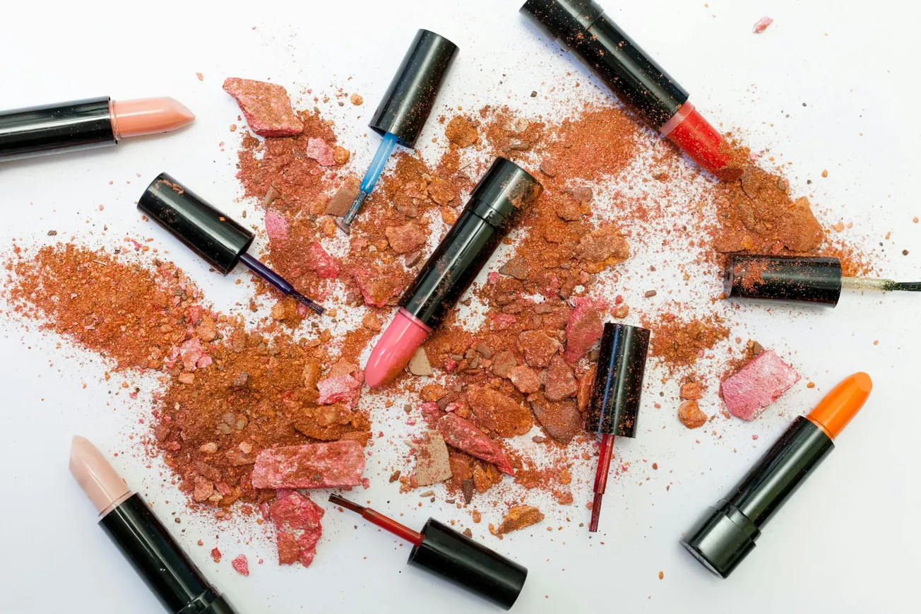 Scattered makeup with broken pink and orange powders, surrounded by various lipstick shades and nail polish bottles.