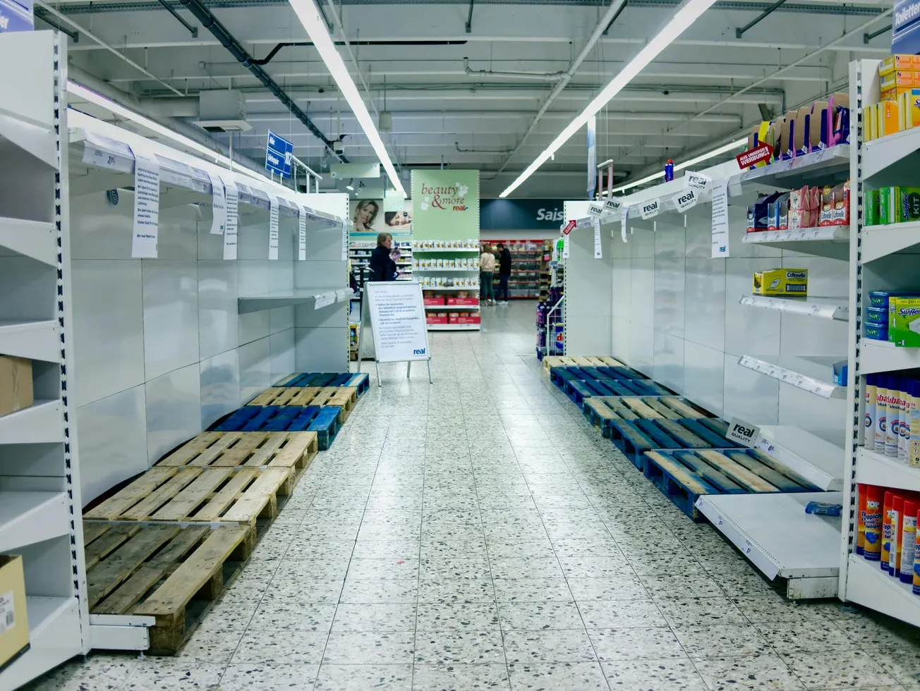 Empty supermarket aisle with bare shelves and wooden pallets. Few items remain on the right. Shoppers in the distance, conveying scarcity.