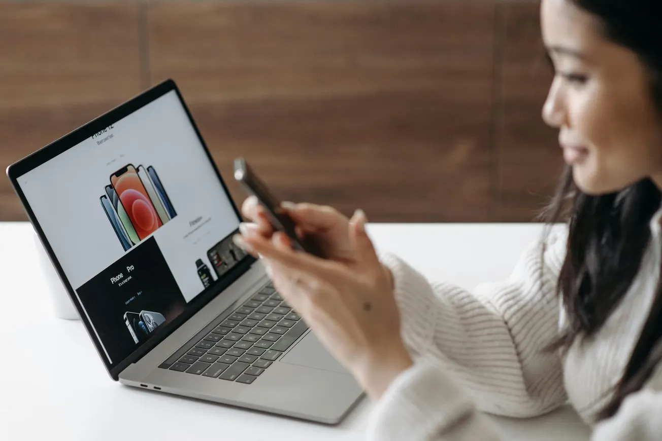A woman in a cozy sweater holds a smartphone, browsing on her laptop displaying a smartphone sales page.