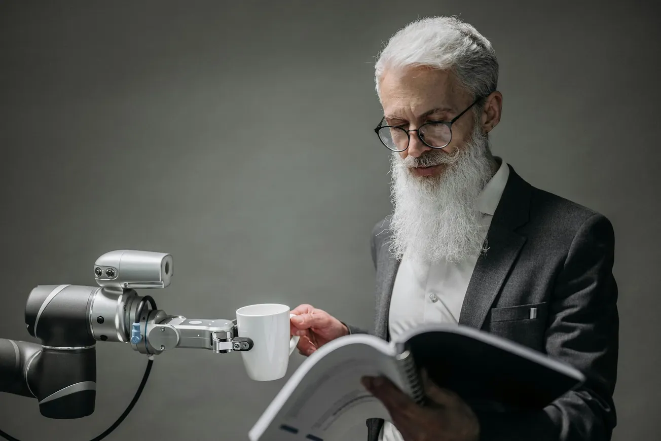 An elderly man with a long white beard and glasses, wearing a suit, reads a book. A robotic arm hands him a coffee mug.