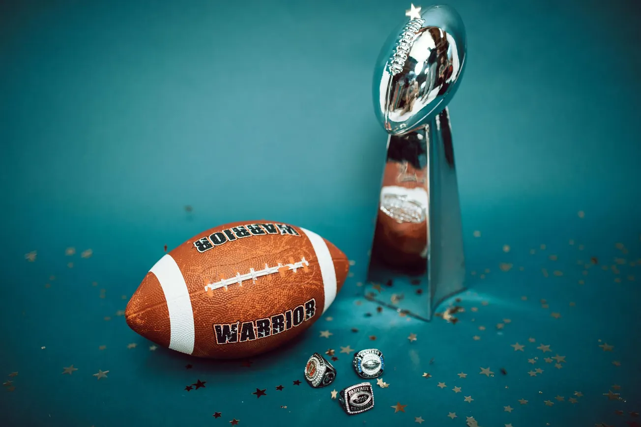 A football and shiny silver trophy are placed on a teal surface, surrounded by black and gold star confetti, alongside three ornate rings.