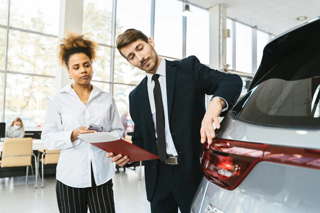 A man in a suit inspects a car's rear light with a thoughtful expression, while a woman beside him holds documents. They are in a bright car showroom.