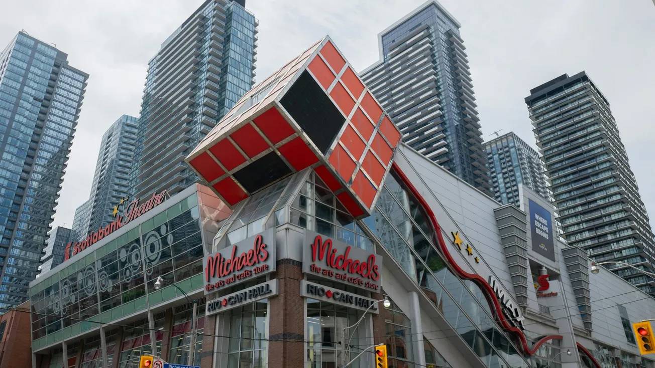 A giant Rubik's Cube structure adorns the front of a modern Michaels building against a backdrop of towering glass skyscrapers under a cloudy sky.