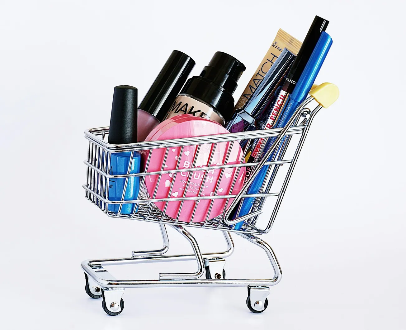 A mini shopping cart filled with various makeup products, including nail polish, blush, mascaras, and lipstick, against a plain white background.