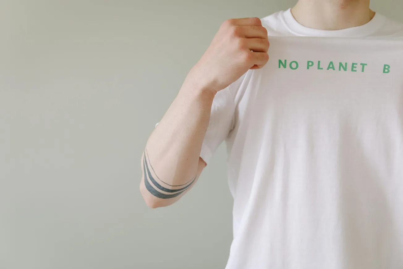 A person wearing a white T-shirt with the text "NO PLANET B" in green. The arm shows a black armband tattoo.