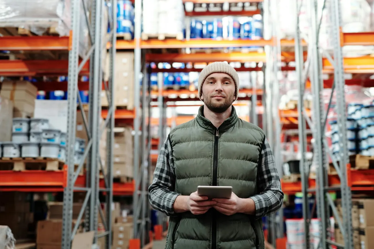 Man in a green vest and beanie holds a tablet, standing in a warehouse with tall shelves filled with boxes and cans.