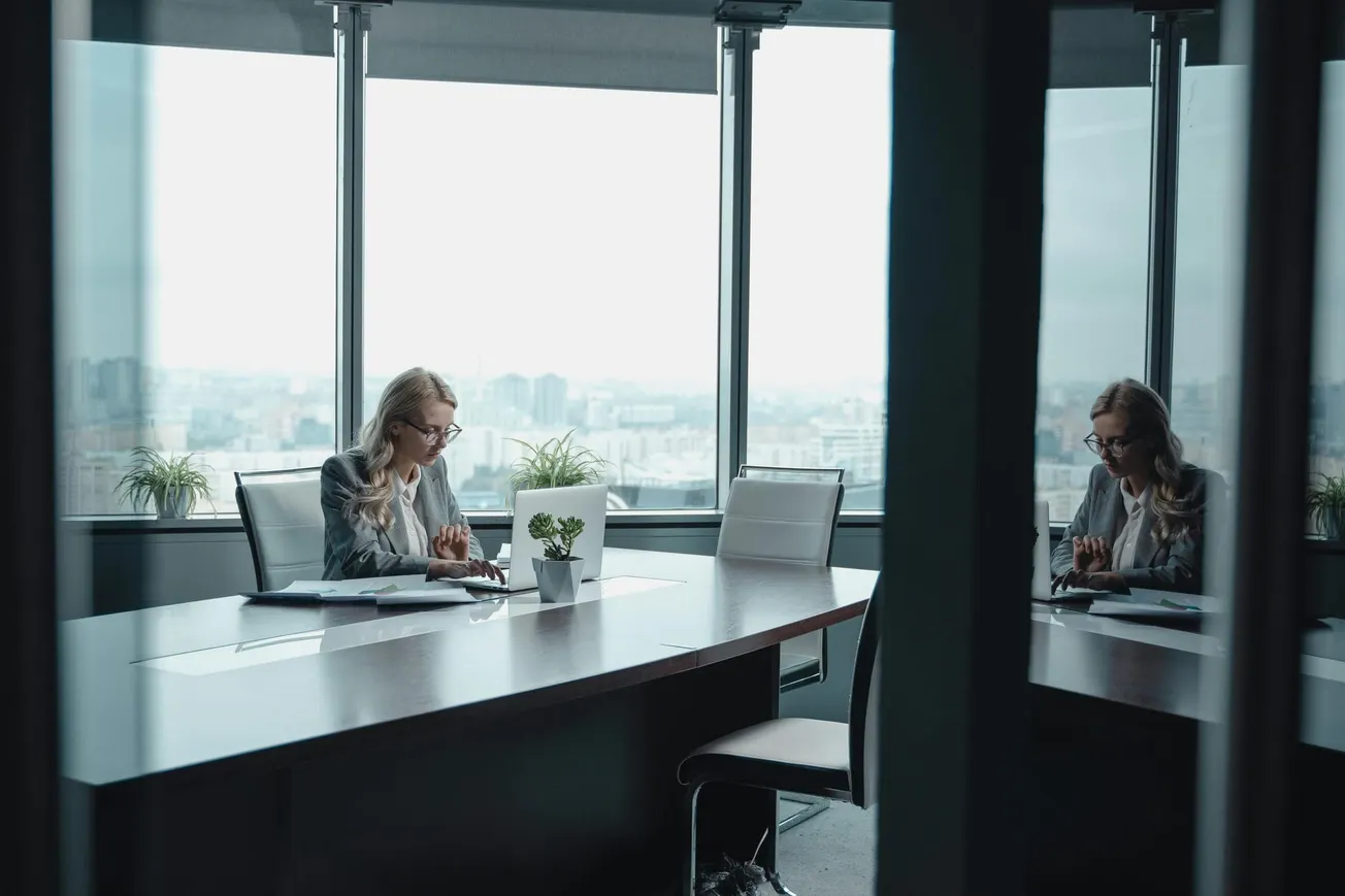 A woman sits at a large conference table in a glass-walled office, working on documents. Outside, the city skyline is visible.