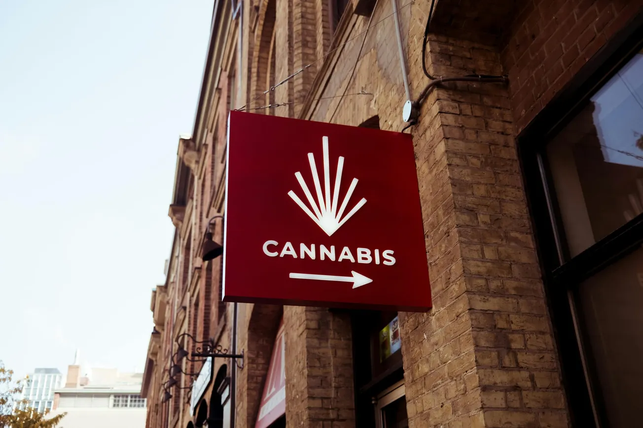 Red sign with white cannabis leaf and text "CANNABIS," featuring an arrow pointing right. Mounted on a brick building with an urban background.