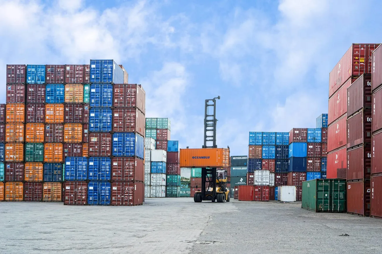 A forklift moves a bright orange shipping container in a port yard, surrounded by colorful stacks of containers under a blue sky with clouds.