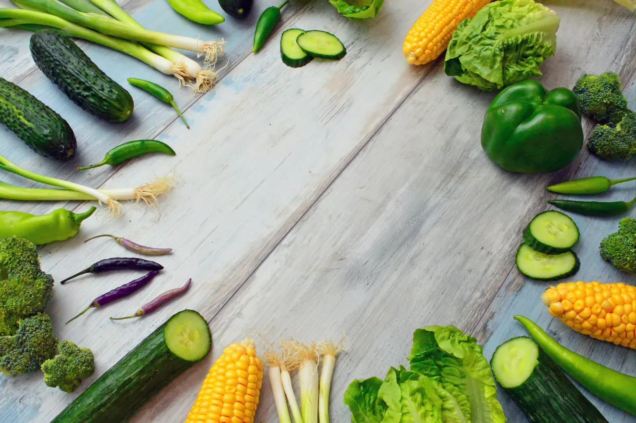 A variety of fresh vegetables are arranged in a semicircle on a light wooden surface. Veggies include cucumbers, green peppers, corn, lettuce, and green onions.