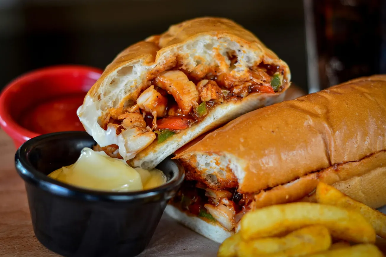 A close-up of a sub sandwich filled with chicken and vegetables, on a wooden board. A red bowl of sauce, black bowl of mayonnaise, and fries are nearby.