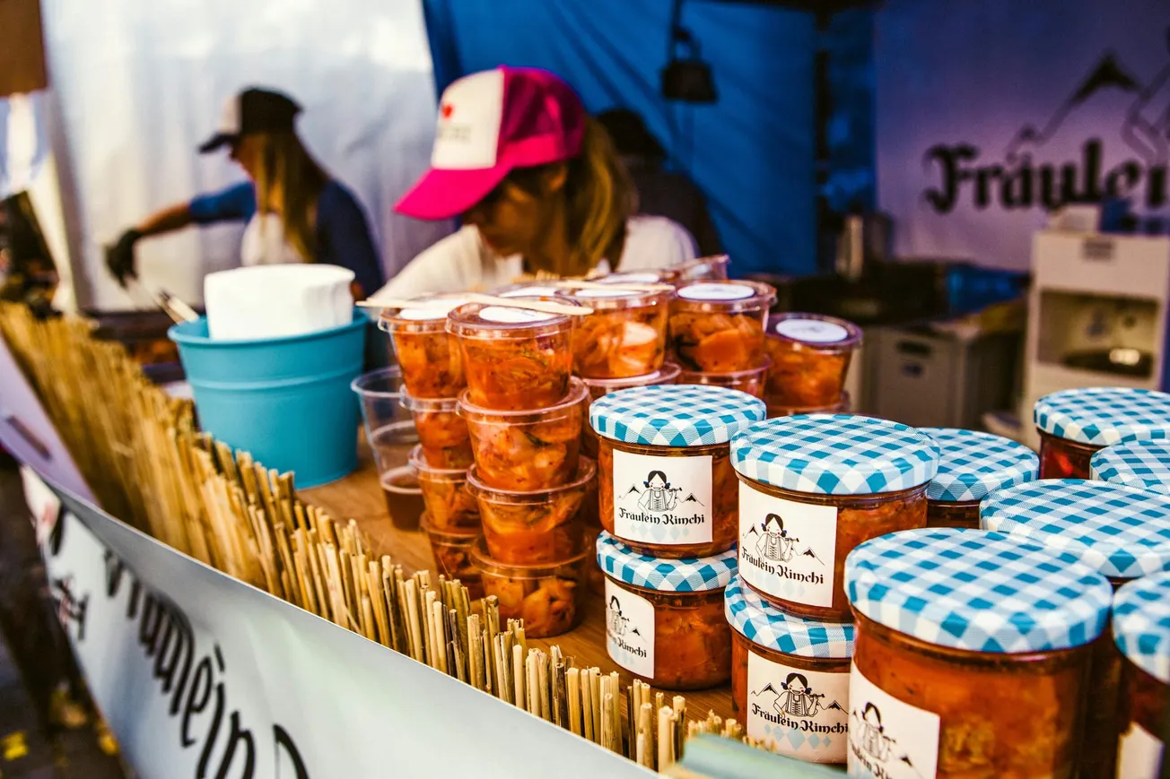 A market stall features stacked jars of kimchi with blue checkered lids. A vendor in a pink cap attends to customers.