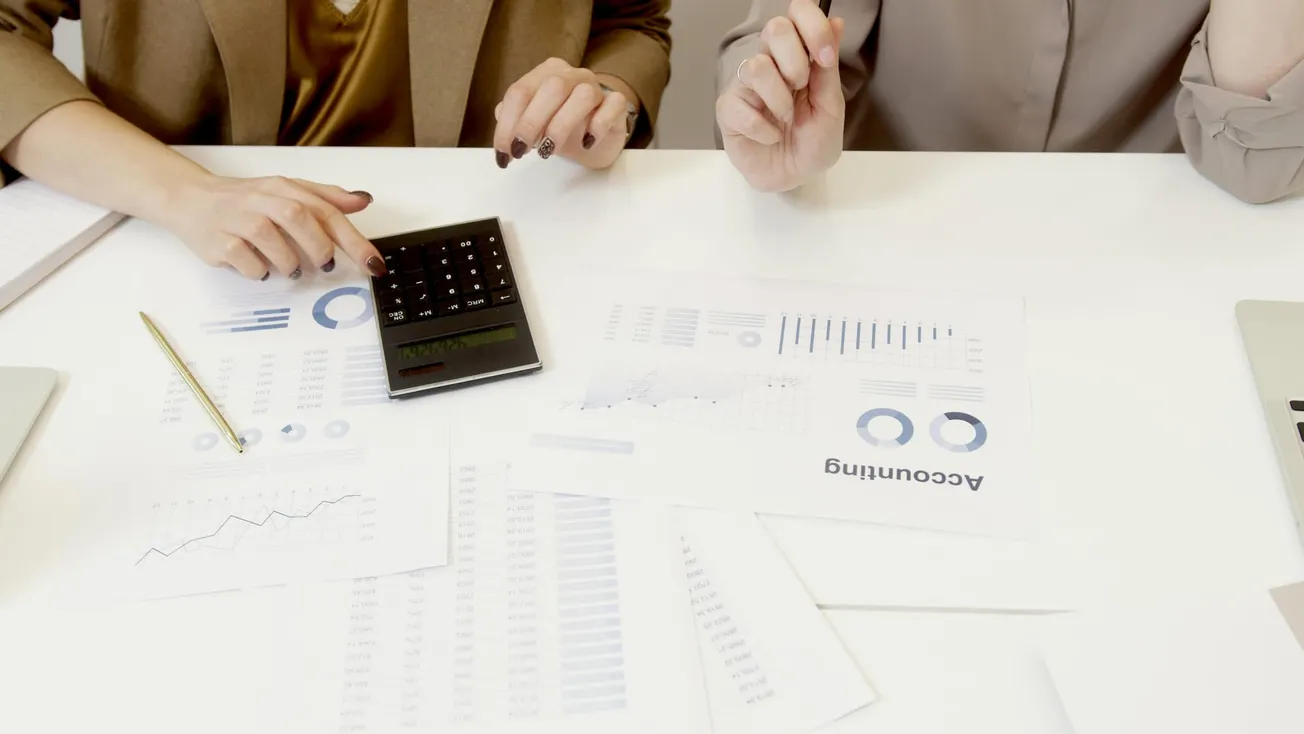 Two individuals working at a desk analyze financial documents. One points to a calculator, surrounded by charts and graphs.