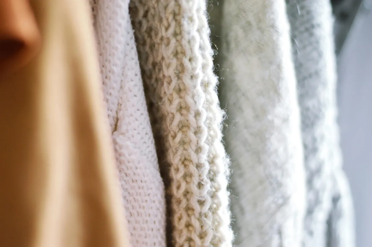 Close-up of various knit sweaters hanging in soft focus. The textures vary, with neutral tones of beige, cream, and light gray, conveying warmth.