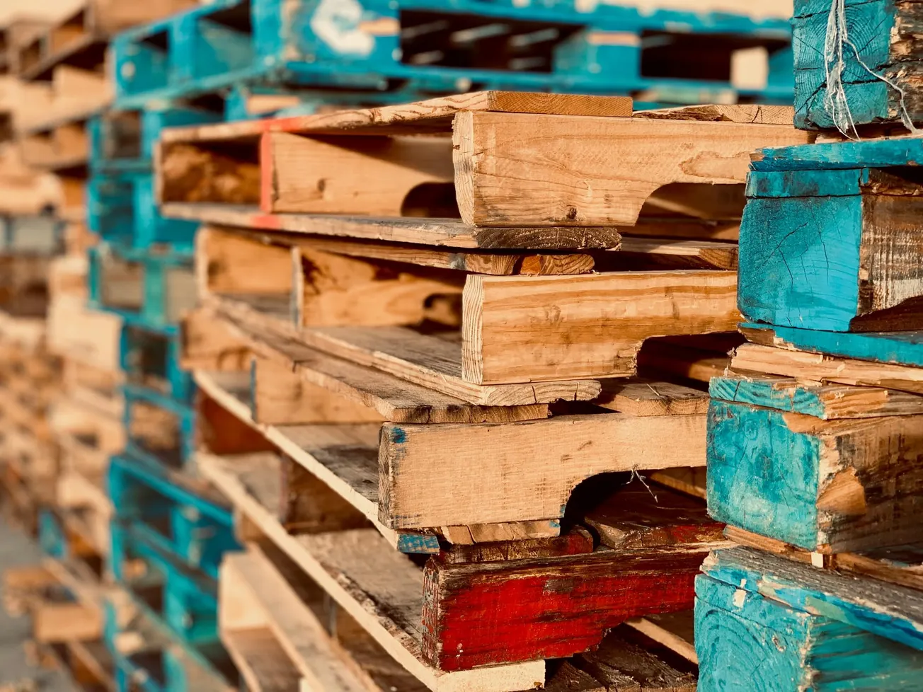 A stack of wooden pallets in varying colors, mainly blue and red, are piled up outdoors. The warm sunlight casts a soft glow, emphasizing their rustic texture.