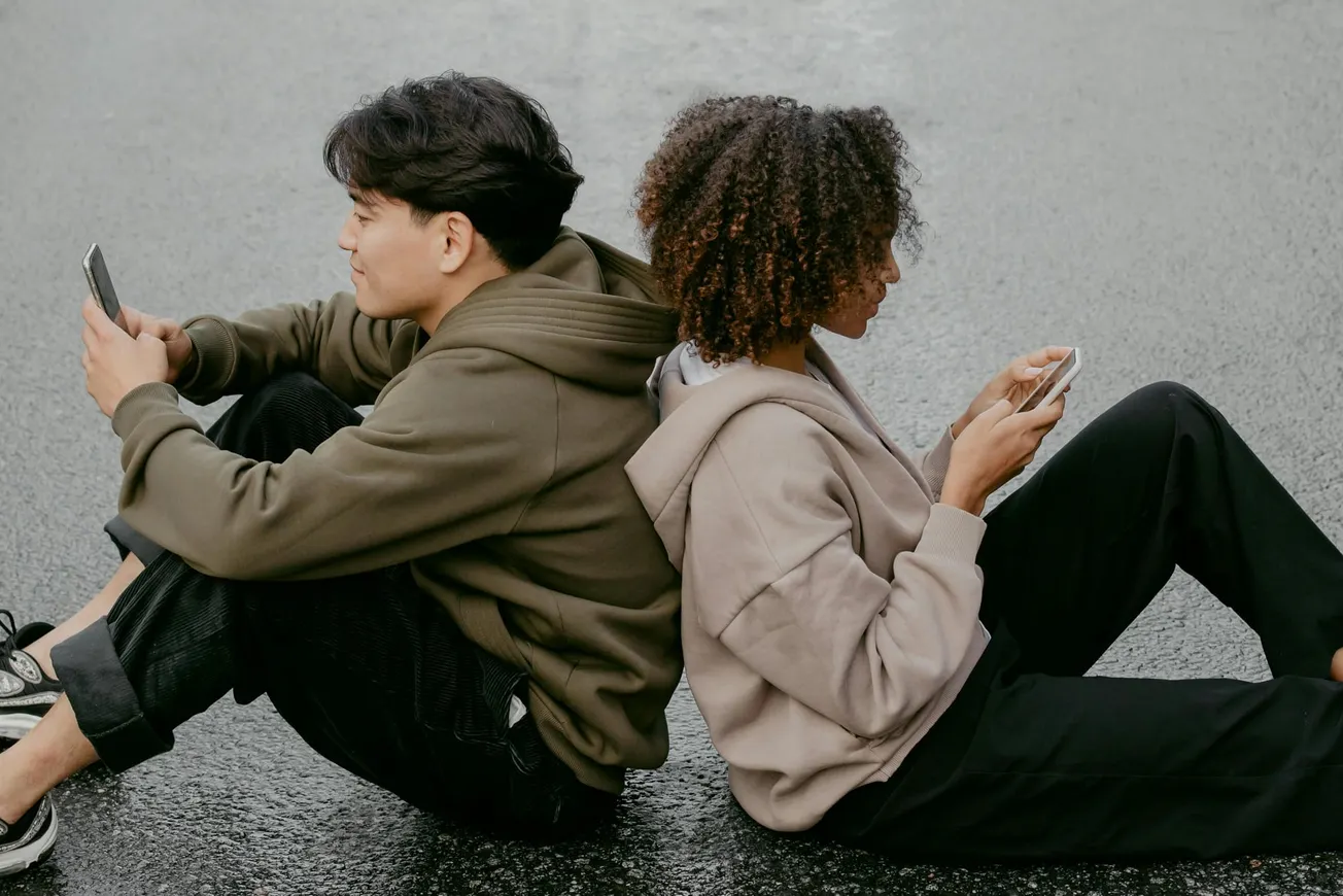 Two people sit back-to-back on a wet pavement, engrossed in their smartphones. Both wear hoodies, creating a relaxed, casual atmosphere.