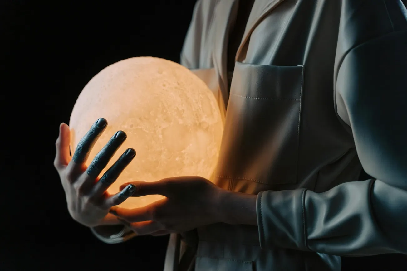 A person in a silky robe holds a glowing moon lamp in their hands. The background is dark, highlighting the warm, serene glow of the lamp.