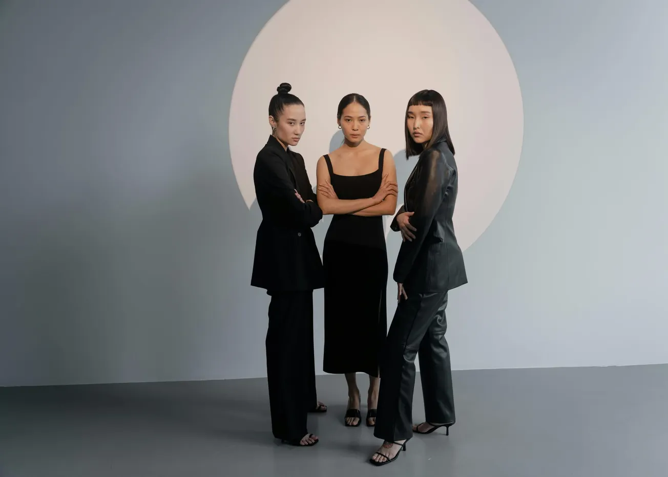 Three women in black outfits stand confidently in front of a large circular light backdrop, exuding strength and unity. Gray setting, modern tone.