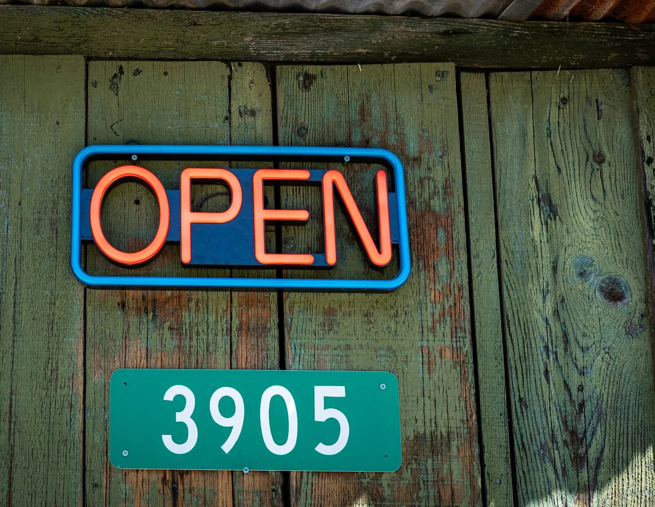 A glowing neon "OPEN" sign is mounted on weathered green wood, above a green sign displaying the number "3905," conveying a welcoming, rustic vibe.