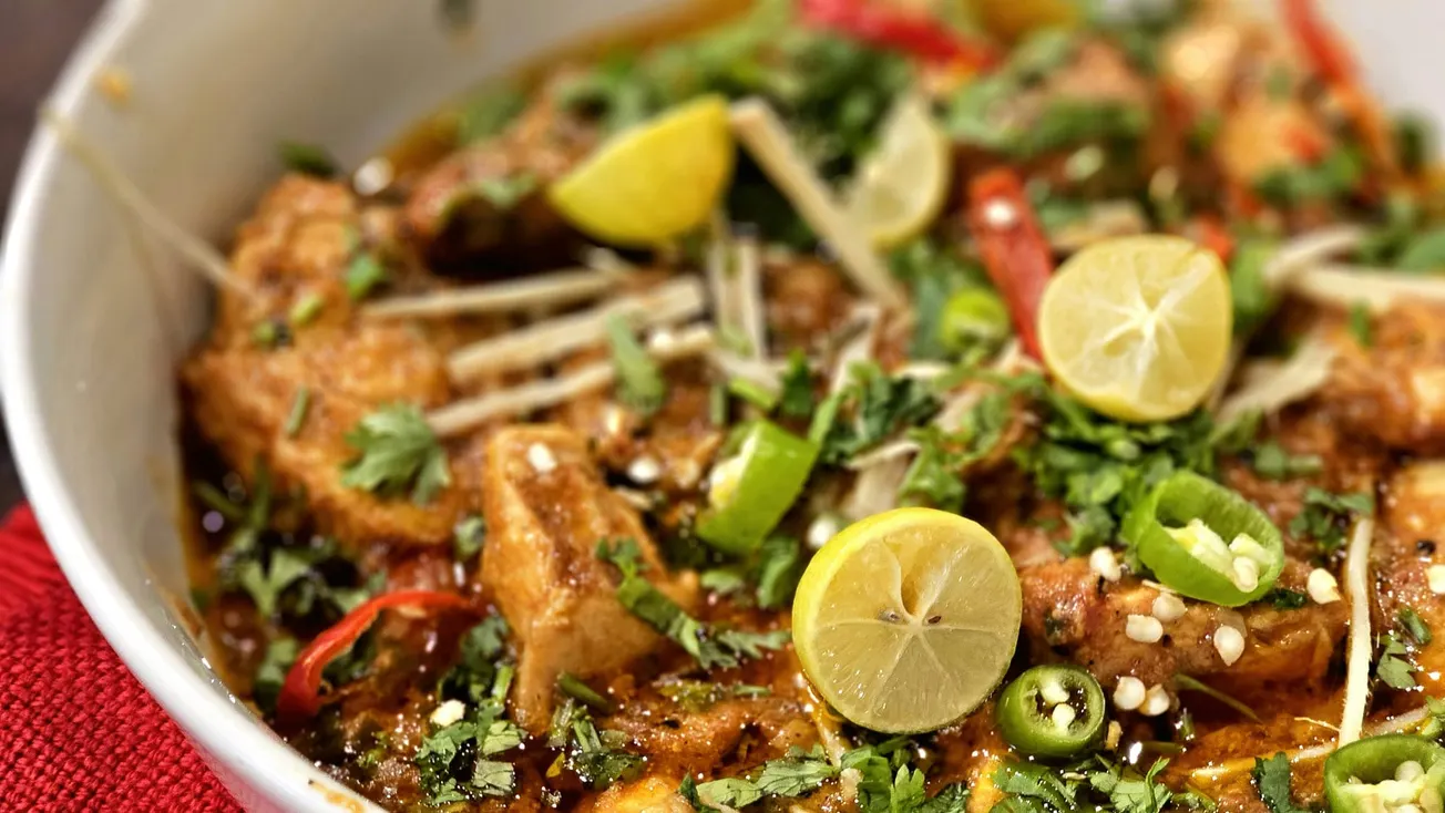A vibrant bowl of chicken curry garnished with halved lime, sliced green and red chilies, fresh cilantro, and ginger strips, exuding a spicy aroma.