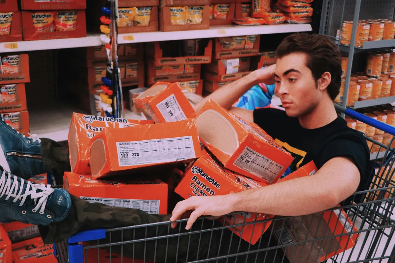 A person sits in a shopping cart filled with orange packages of ramen noodles in a grocery store aisle.