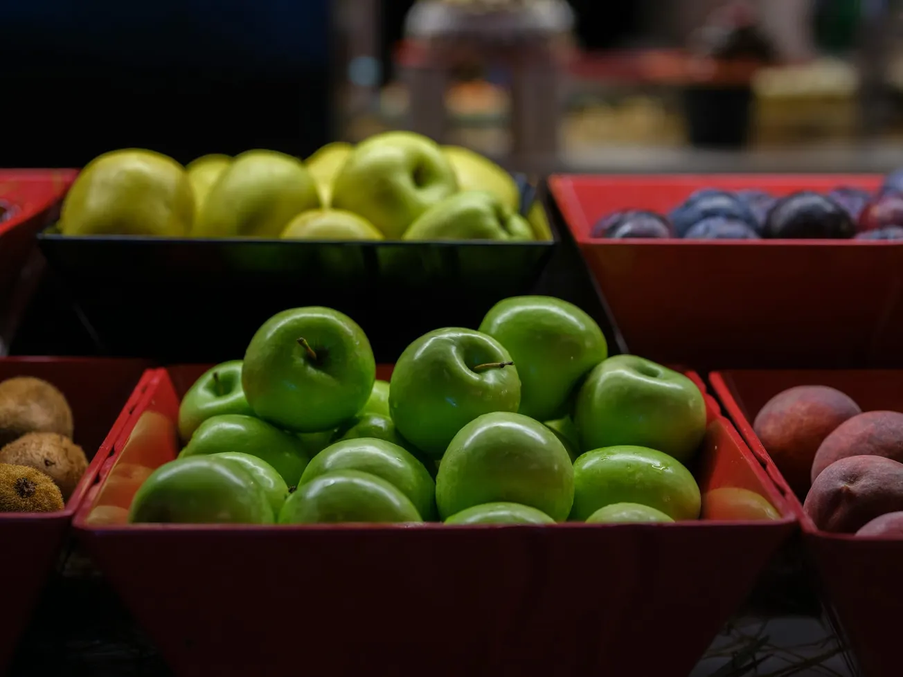 Close-up of fresh green apples in a red tray, surrounded by various fruits like yellow apples, plums, and peaches in a dimly lit market setting.