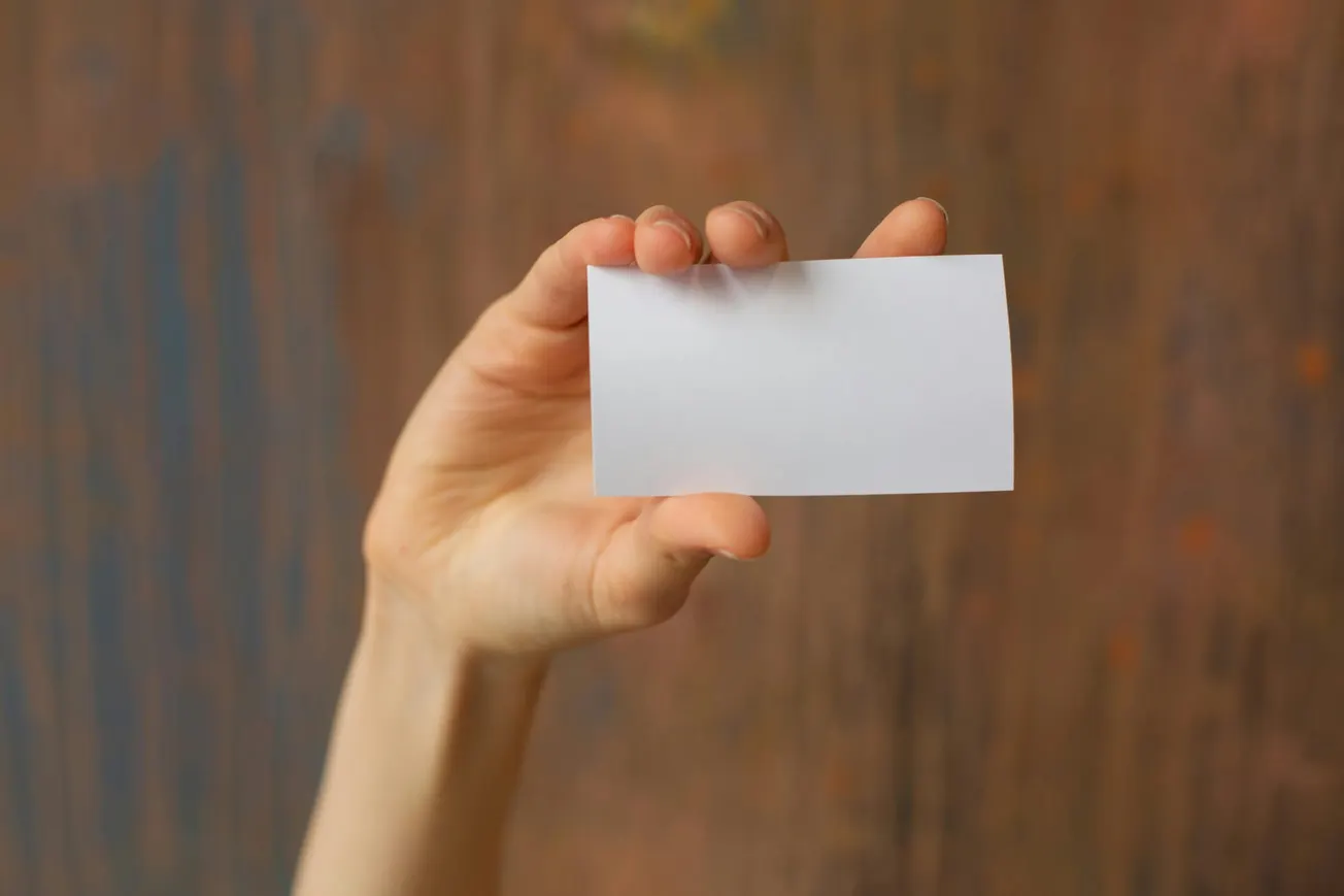 A hand holds a blank white card against a textured brown background.