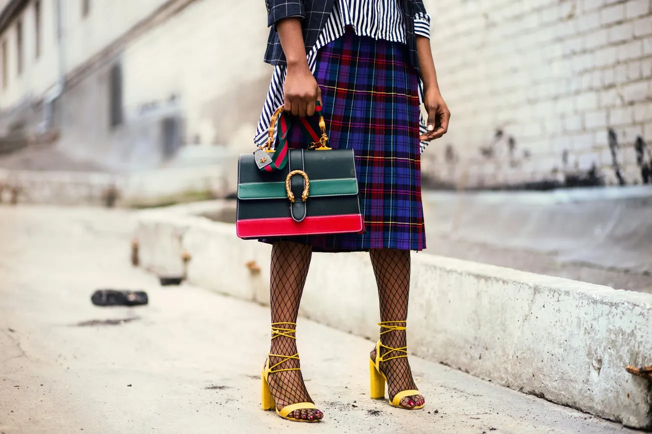 Fashionable street style: person in a checkered skirt and striped shirt holds a designer handbag. Yellow lace-up heels and fishnet tights add flair. Urban setting.