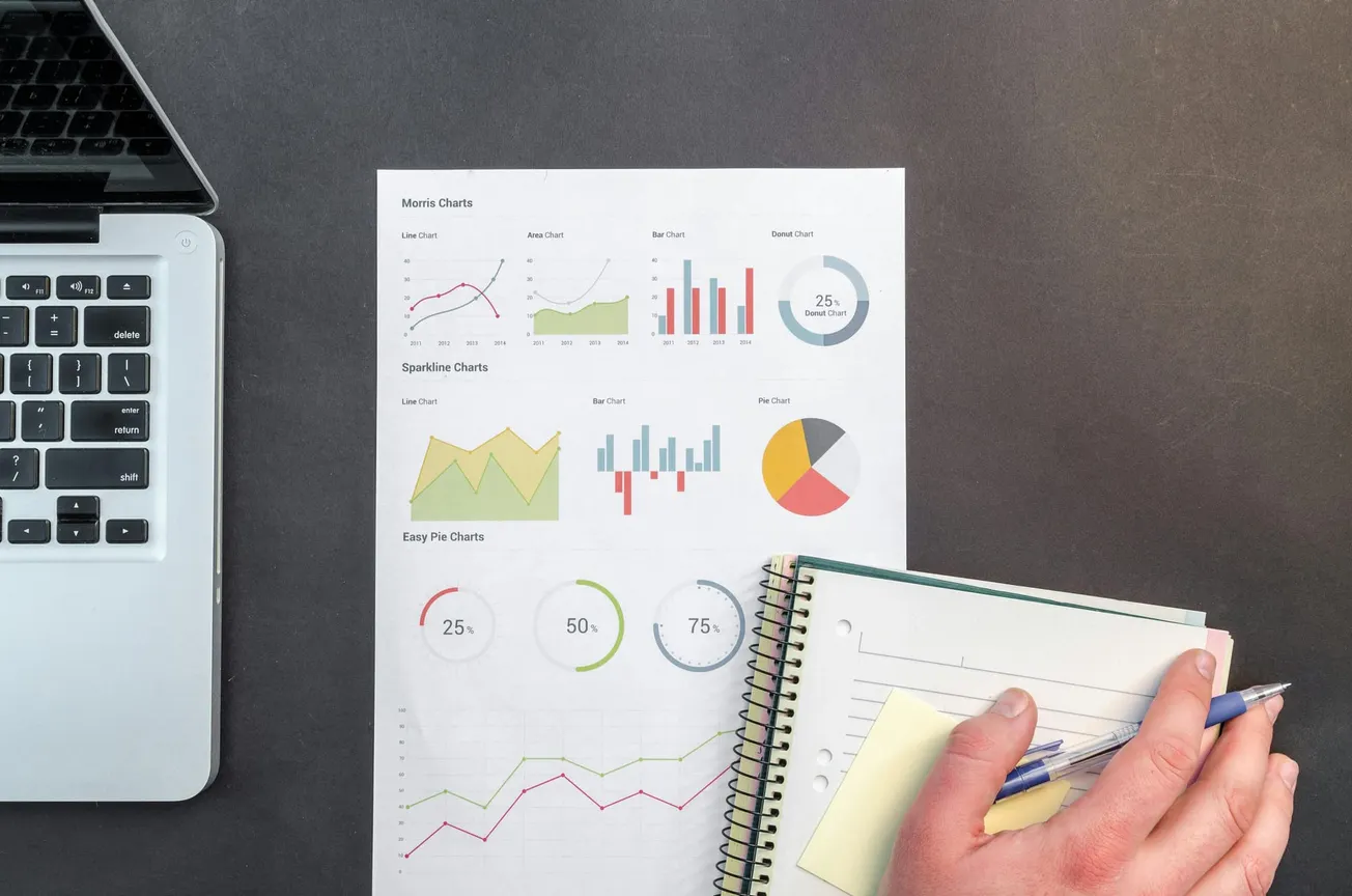 A laptop and a paper with colorful charts and graphs are on a gray desk. A hand holding a pen is poised over a spiral notebook, suggesting a work-focused setting.