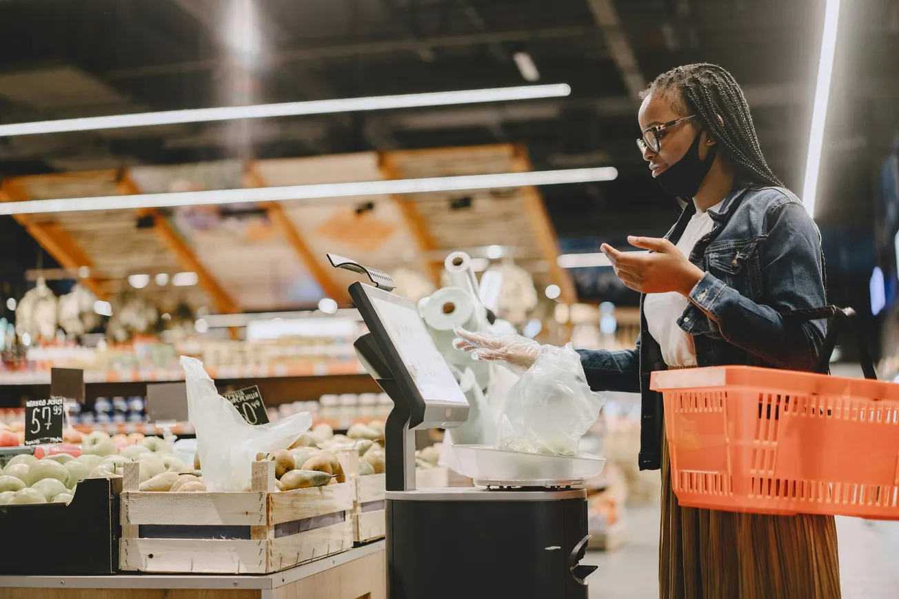 A woman in a denim jacket weighs produce at a self-checkout in a supermarket. She holds a red basket and wears a mask under her chin.
