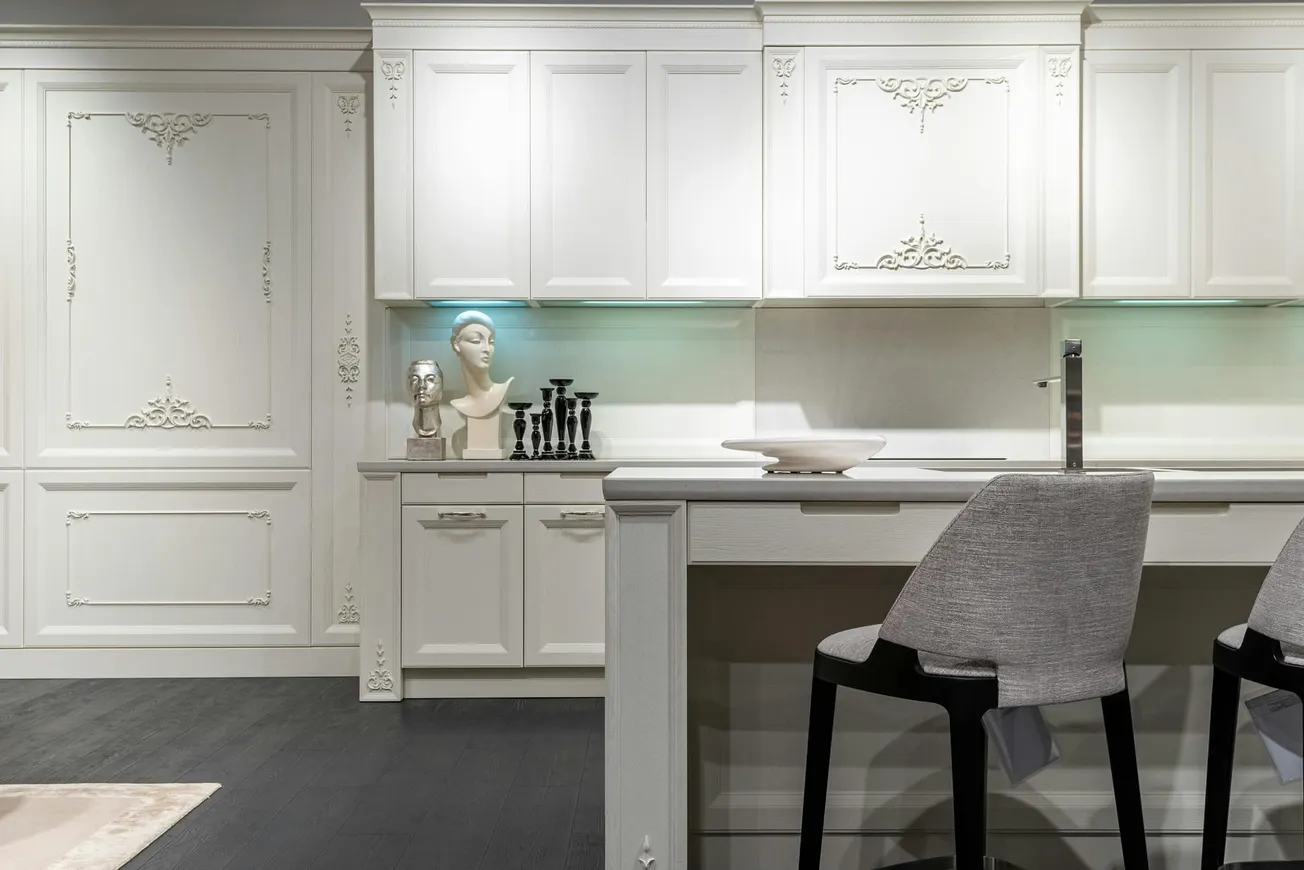 Elegant white kitchen with ornate cabinets, black candlesticks, and a marble bust on the counter. Two upholstered chairs at a sleek island.