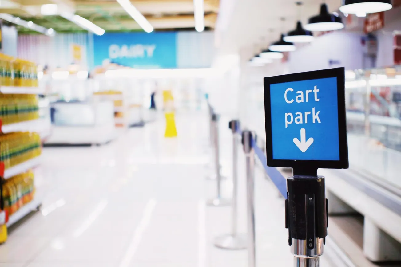 A supermarket aisle with a blue "Cart park" sign in focus on the right, and blurred shelves of products under bright lights in the background.