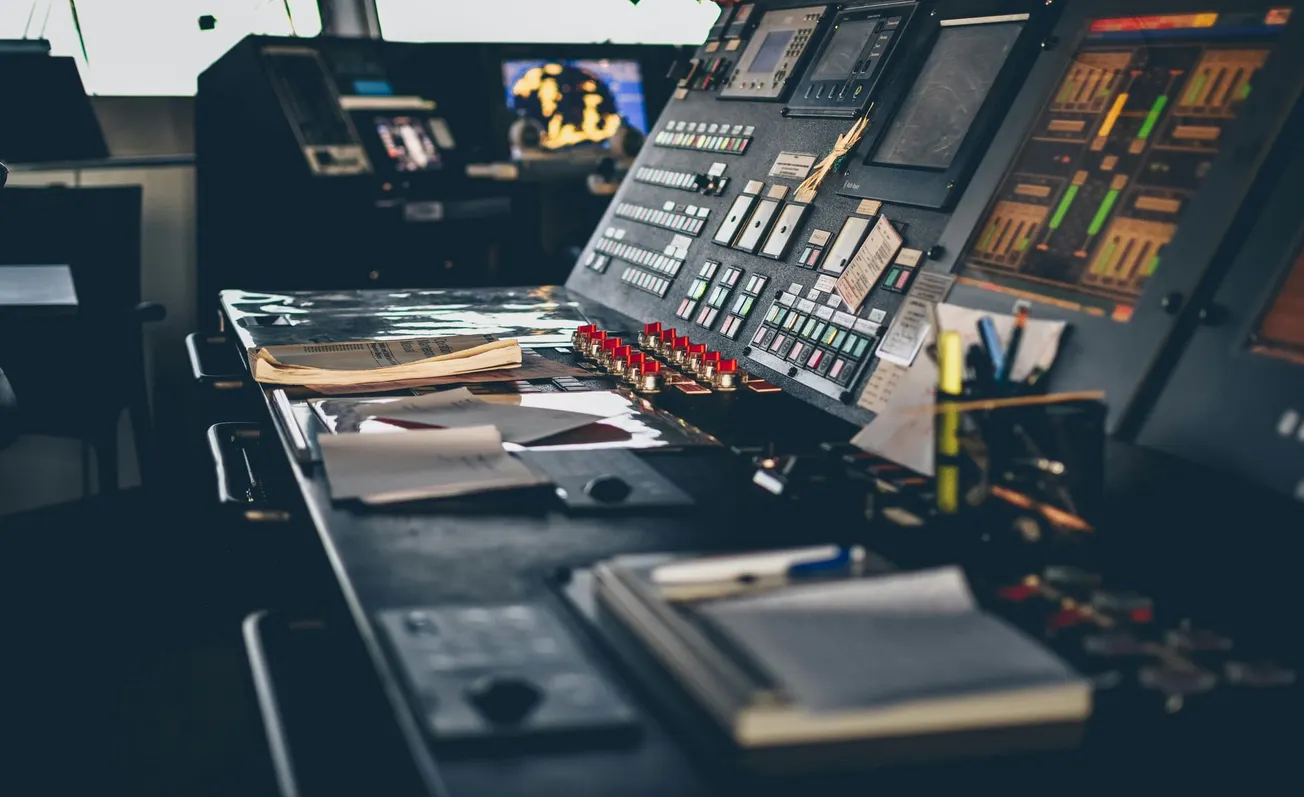 A dimly lit control room with a large panel covered in switches, buttons, and screens. Papers and notebooks are scattered on the desk.