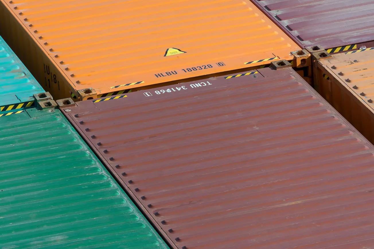 Stacked shipping containers in green, orange, and brown, viewed from above. The containers display visible ID numbers and black and yellow caution markings.