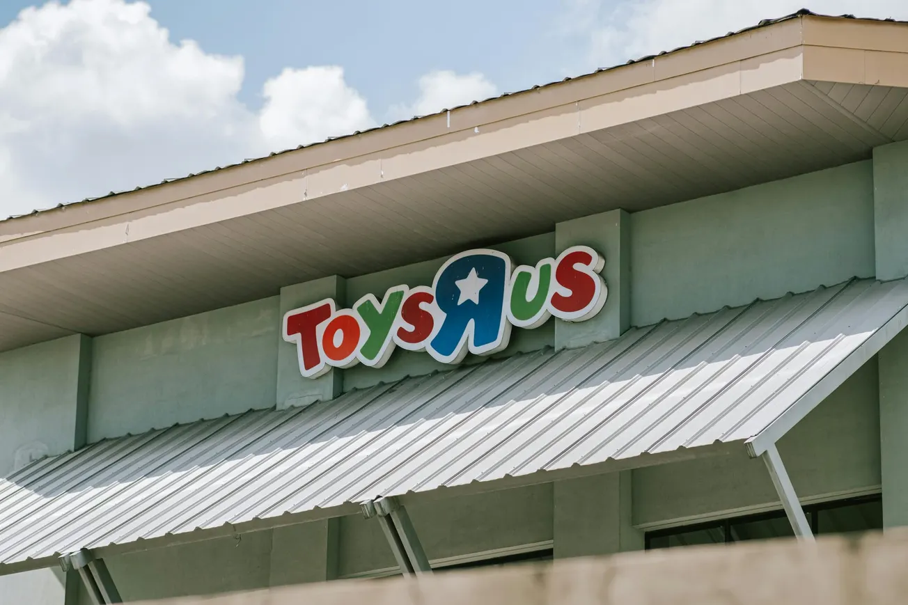 Signage of a Toys R Us store on a building with a gray roof. The colorful logo appears against a light green wall under a partly cloudy sky.
