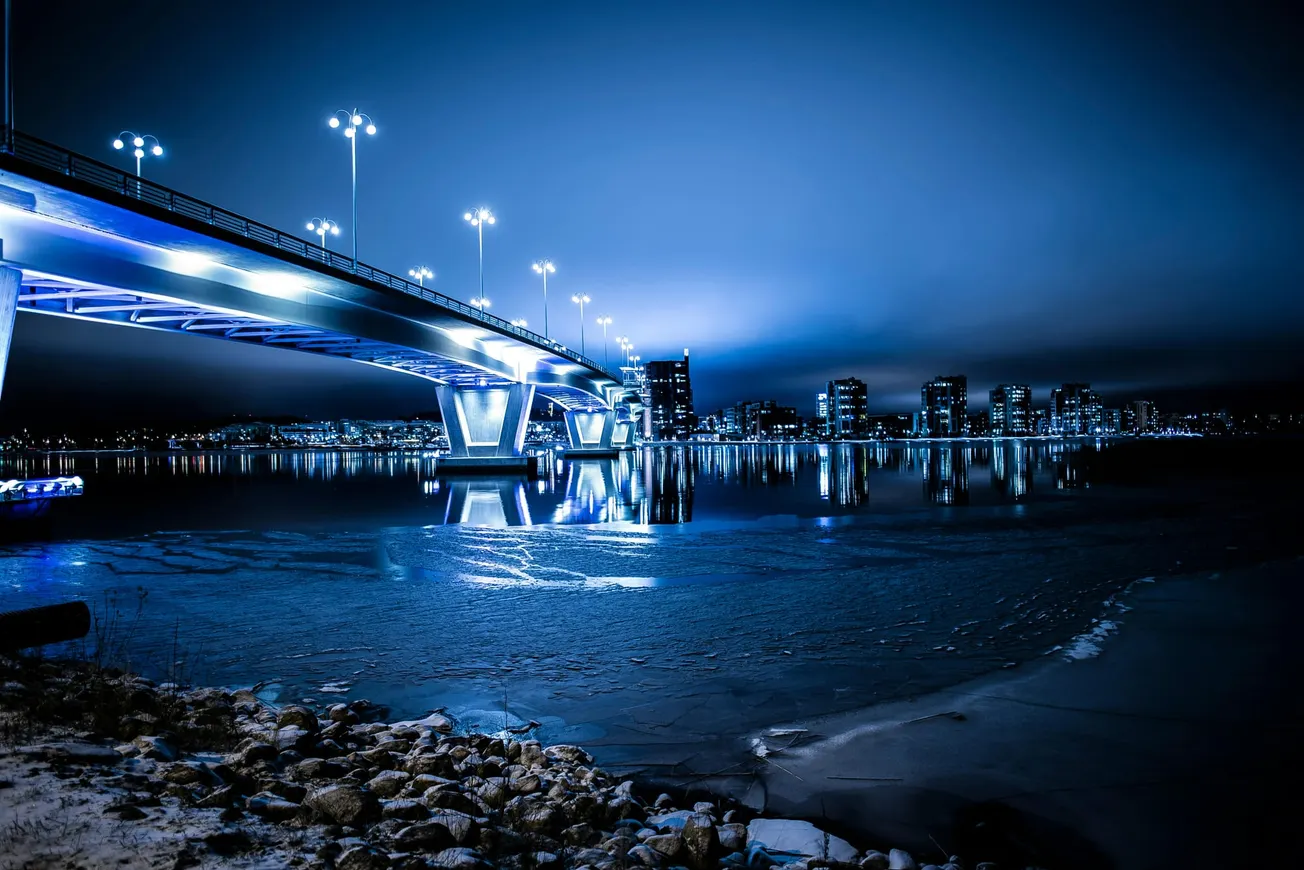 Illuminated bridge reflecting on a calm river at night, with city skyline in the background. The scene conveys a serene, futuristic atmosphere.