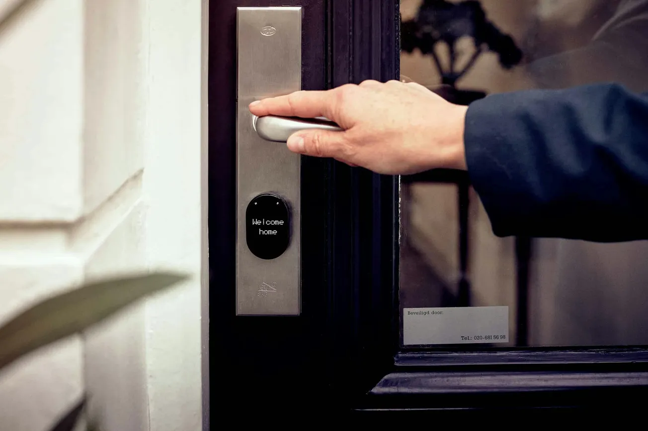 A hand in a dark sleeve reaches for a modern door handle on a sleek metal keypad lock displaying the message "Welcome home."
