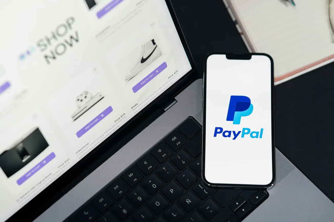 Laptop with online shopping site and "Shop Now" text on screen, next to a smartphone displaying the PayPal logo. Represents digital shopping and payments.
