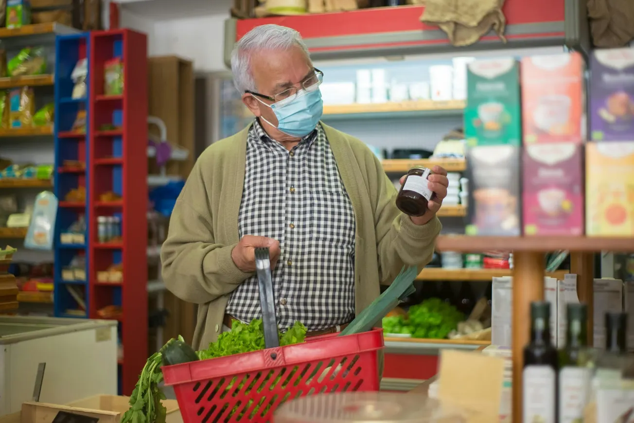 Elderly man in a mask holds a jar while shopping in a grocery store. He has a basket with vegetables, standing near colorful box displays.