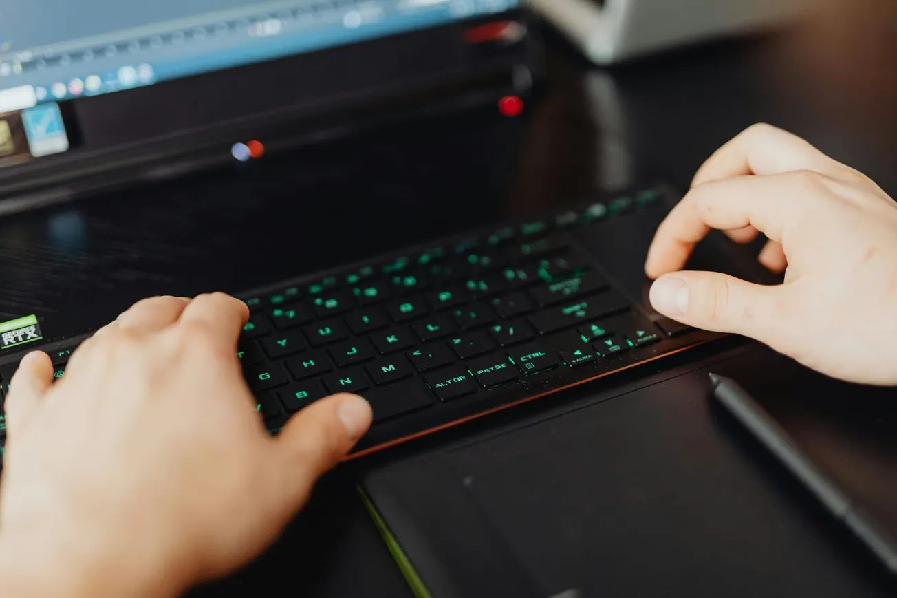 Hands typing on a keyboard with green backlighting. A laptop screen is visible, suggesting a focused, tech-driven work environment.