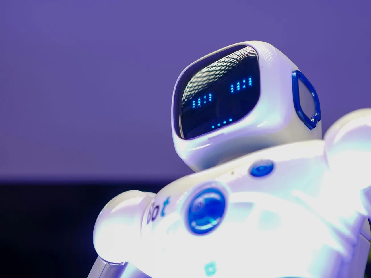 A white robot with a glossy finish is shown against a purple backdrop. Its screen face displays blue lights resembling eyes. The mood is futuristic and friendly.