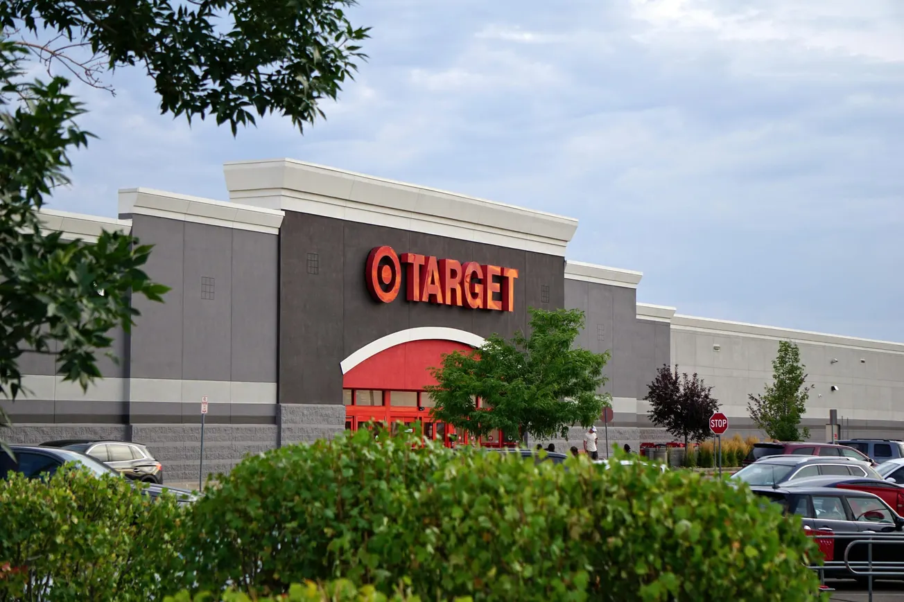 The image shows a Target store exterior with red signage against a gray building. Cars are parked in front, surrounded by green trees under a cloudy sky.