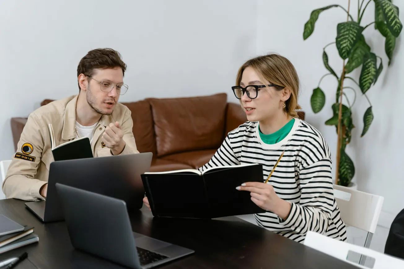 A man and woman sit at a table with laptops, discussing notes in a book. The setting is a cozy office with a plant and a brown couch, conveying collaboration.