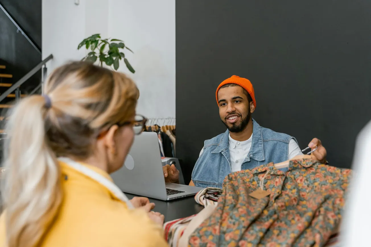 A man in a denim vest and orange beanie smiles while showing a floral dress to a woman in a yellow sweater at a boutique counter with a laptop.