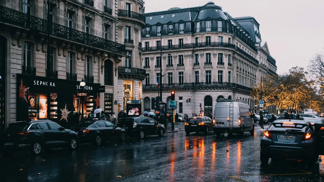 A busy Paris street on a rainy day, with reflections on the wet pavement. Cars and a white van move near elegant historic buildings and lit-up storefronts.