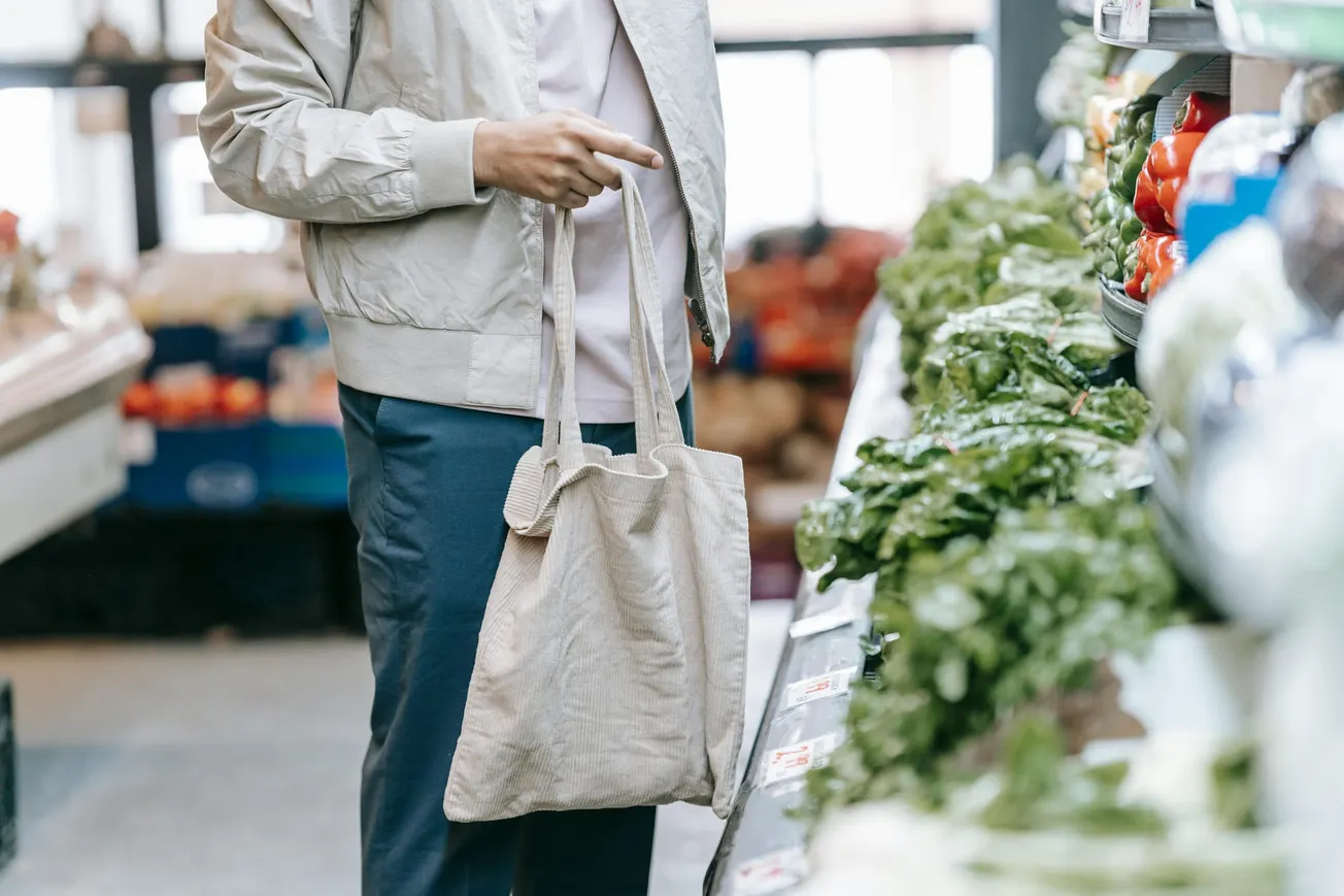 Person in a beige jacket shopping for leafy greens in a grocery store. They hold a reusable bag, creating an eco-friendly shopping vibe.
